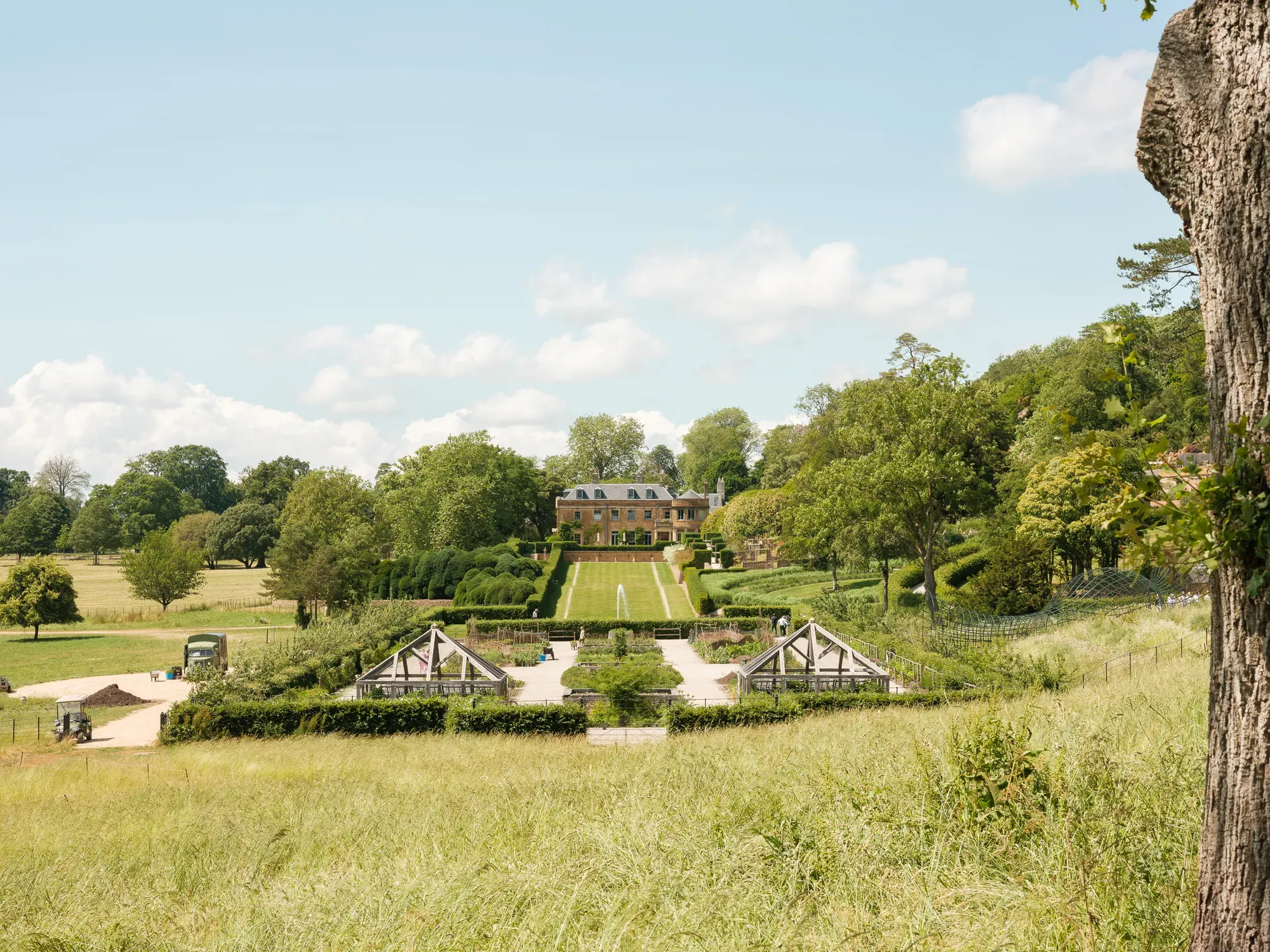 Formal garden and country house viewed across meadow under blue sky with scattered clouds.