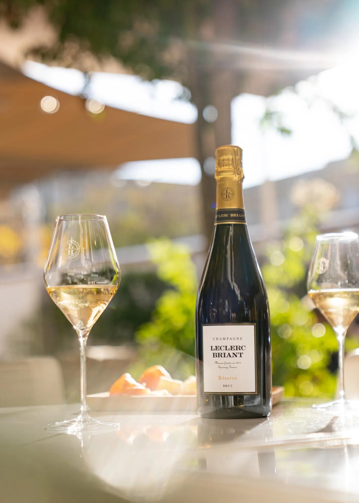 Bottle of Leclerc Briant Champagne between two filled glasses on a sunlit terrace table.