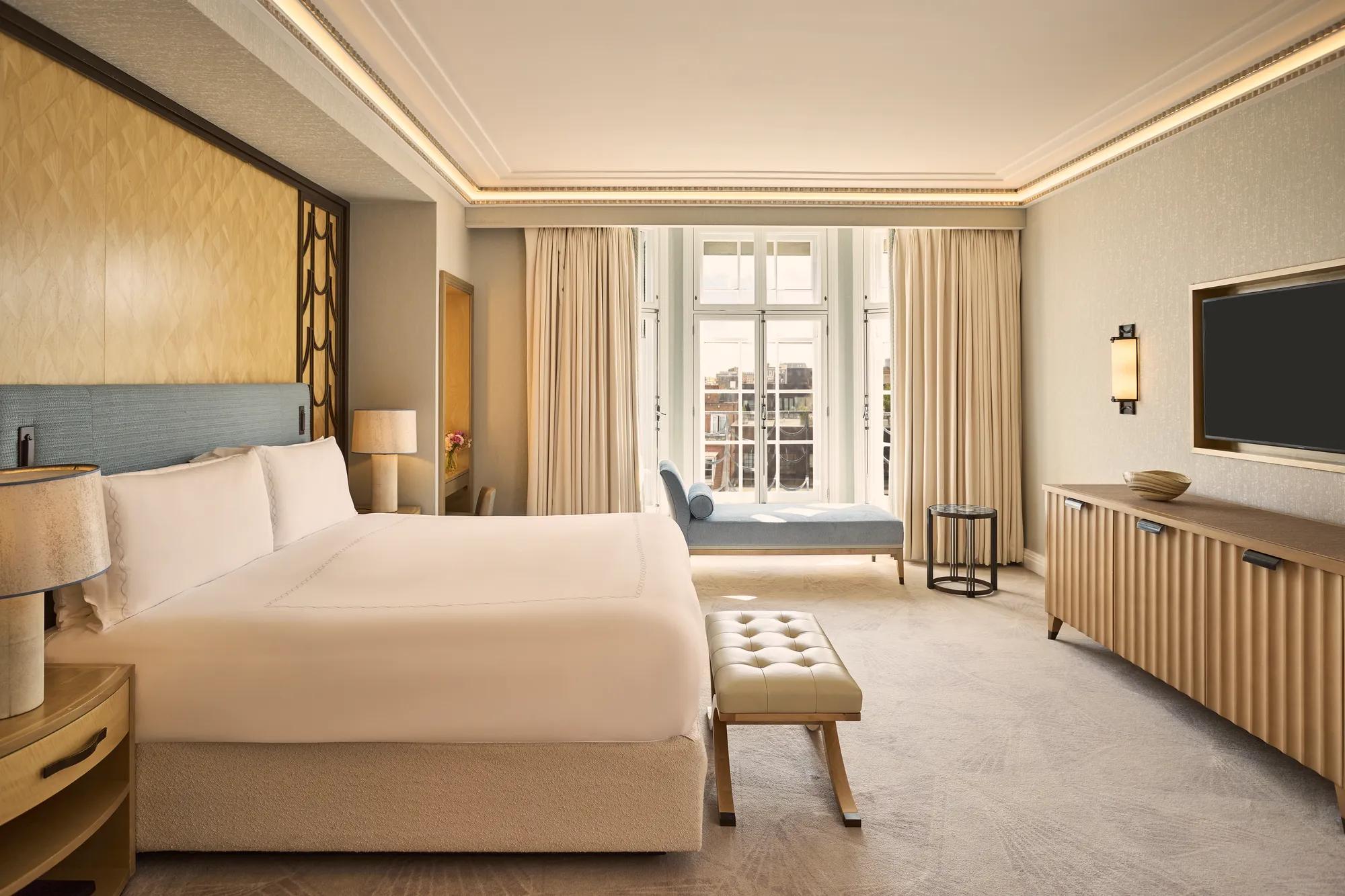Contemporary bedroom with a king-size bed, upholstered bench, wall-mounted television, and chaise longue positioned beside floor-to-ceiling windows.
