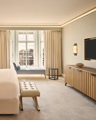 Contemporary bedroom with a king-size bed, upholstered bench, wall-mounted television, and chaise longue positioned beside floor-to-ceiling windows.