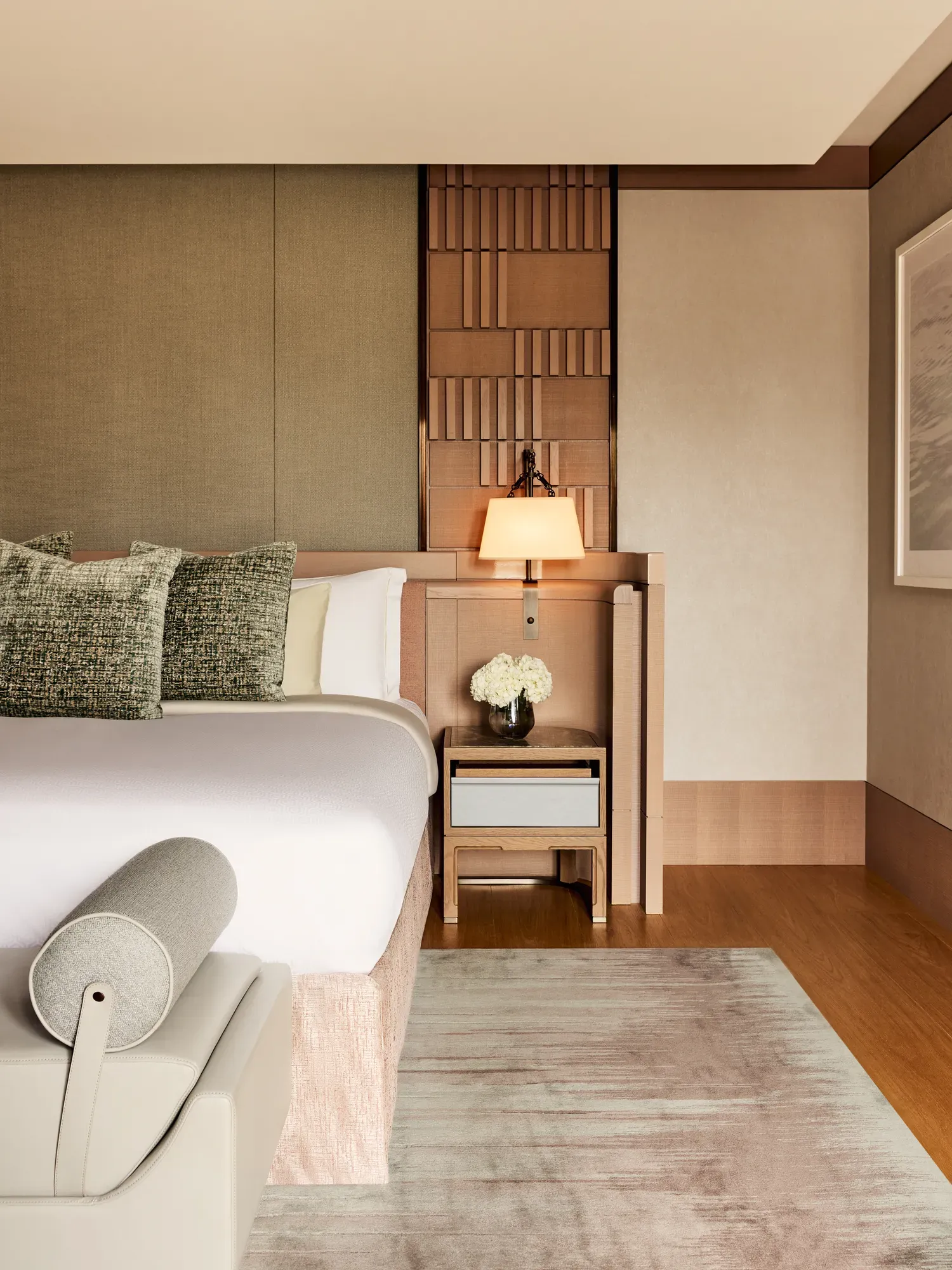 Luxurious bedroom corner with upholstered bed, patterned cushions, bedside table, wall lamp, and neutral-toned textured wall panels.