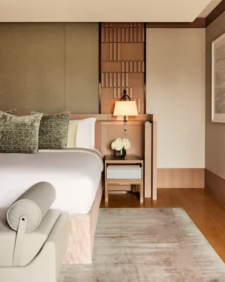 Luxurious bedroom corner with upholstered bed, patterned cushions, bedside table, wall lamp, and neutral-toned textured wall panels.