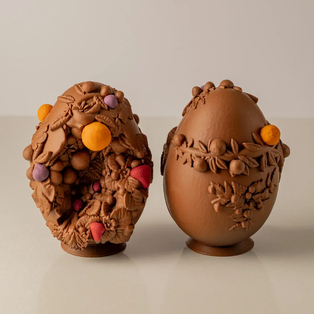 Two decorative chocolate Easter eggs with sculpted leaves, berries, and fruit details displayed on small stands against neutral background.