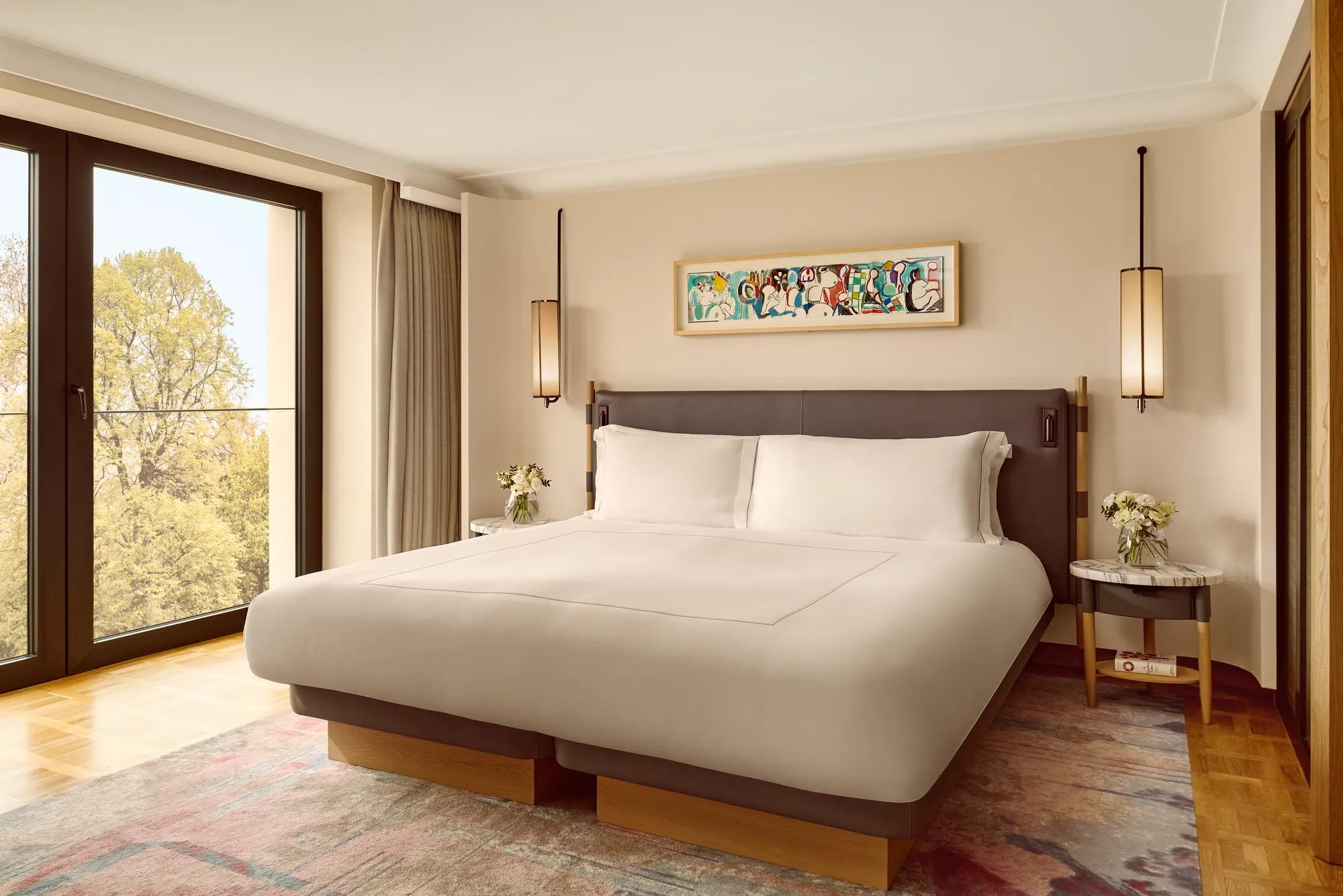 Elegant bedroom with a king bed, modern pendant lights, and a colorful abstract artwork above the headboard, framed by full-height windows overlooking greenery.