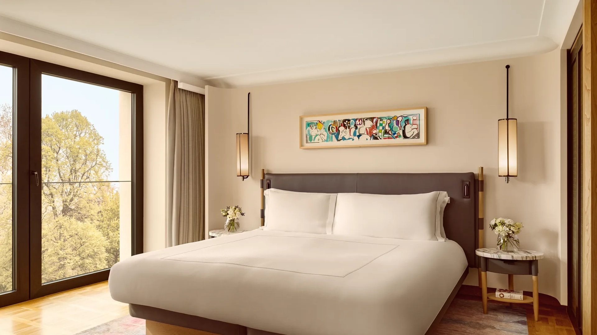 Elegant bedroom with a king bed, modern pendant lights, and a colorful abstract artwork above the headboard, framed by full-height windows overlooking greenery.