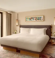 Elegant bedroom with a king bed, modern pendant lights, and a colorful abstract artwork above the headboard, framed by full-height windows overlooking greenery.