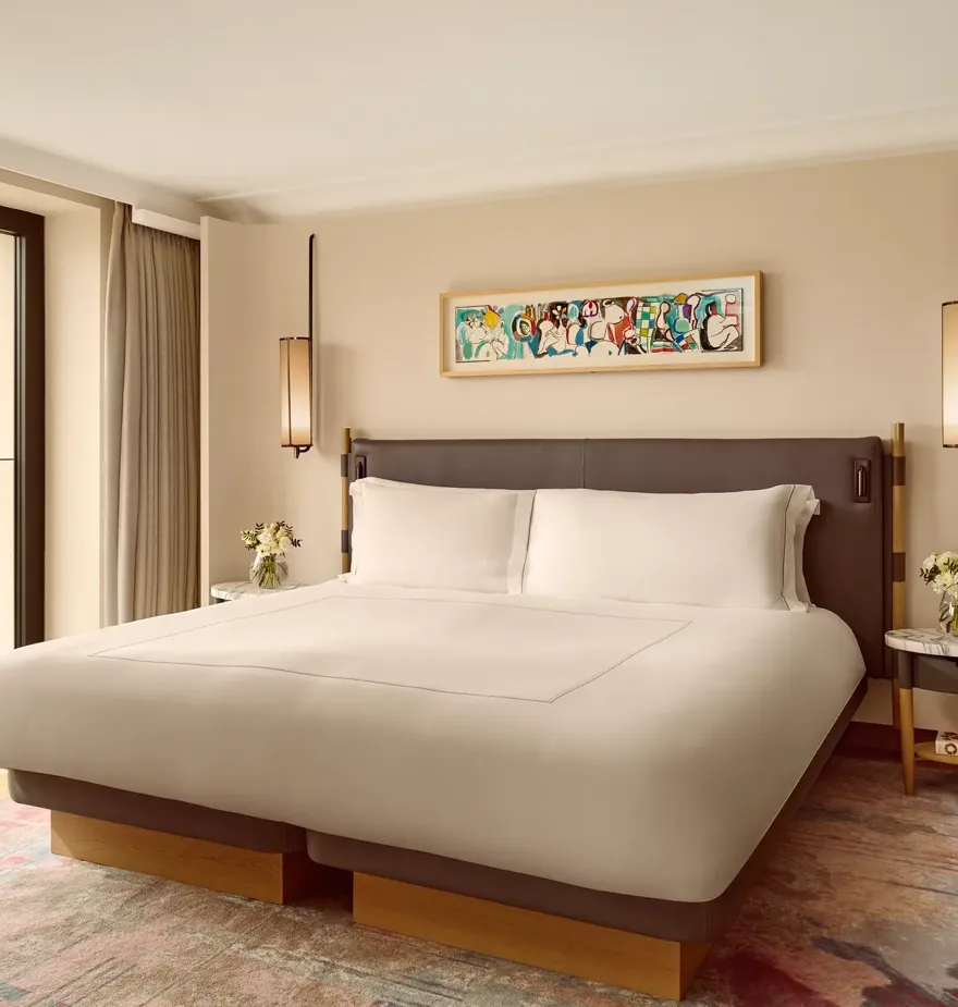 Elegant bedroom with a king bed, modern pendant lights, and a colorful abstract artwork above the headboard, framed by full-height windows overlooking greenery.