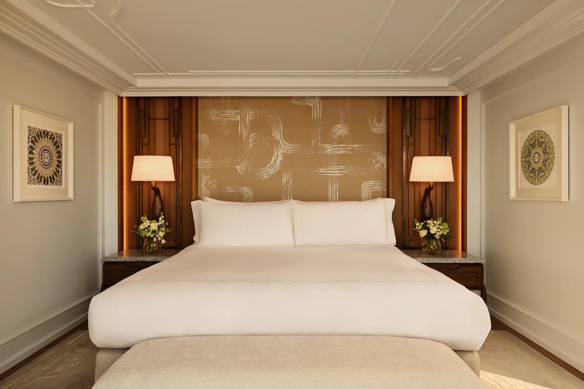 Elegant bedroom with a king-size bed dressed in white linens, framed by warm wooden panels, patterned gold feature wall, bedside tables with matching lamps, and floral arrangements on each side.