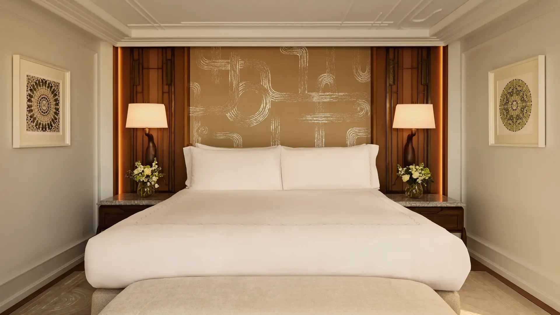 Elegant bedroom with a king-size bed dressed in white linens, framed by warm wooden panels, patterned gold feature wall, bedside tables with matching lamps, and floral arrangements on each side.