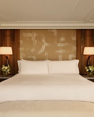 Elegant bedroom with a king-size bed dressed in white linens, framed by warm wooden panels, patterned gold feature wall, bedside tables with matching lamps, and floral arrangements on each side.