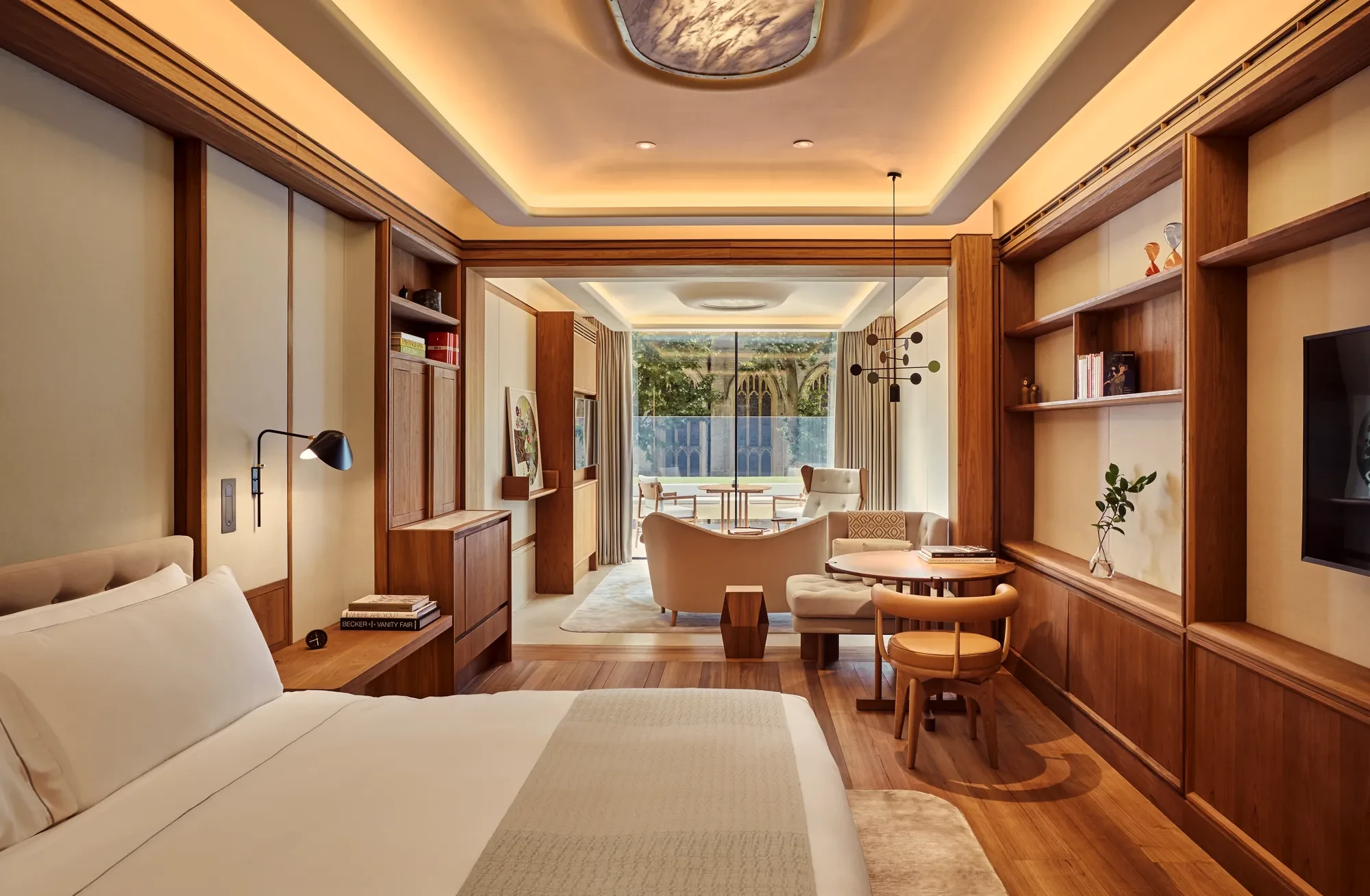 Warm wood-panelled suite with bed, desk and shelving, opening to a sitting area and balcony overlooking gardens.