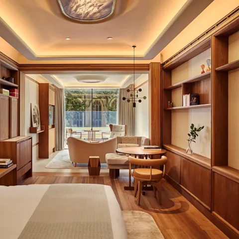 Warm wood-panelled suite with bed, desk and shelving, opening to a sitting area and balcony overlooking gardens.