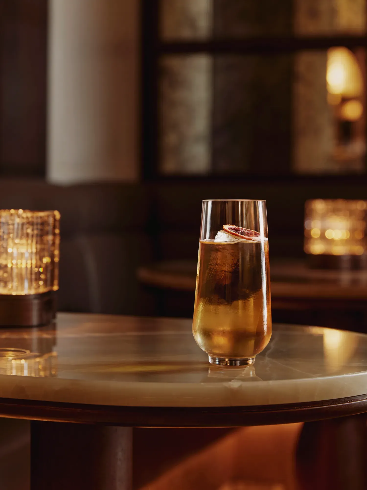 Tall highball with clear ice and citrus slice on a marble table in a dim lounge.