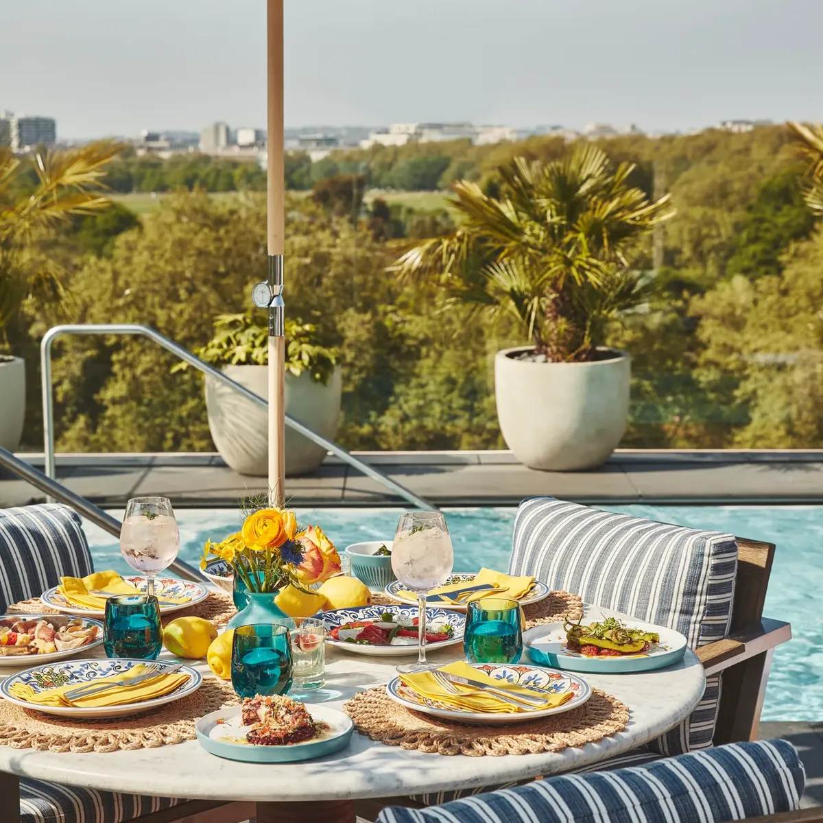 Poolside dining table with striped chairs, colourful plates, and yellow napkins under blue-and-white umbrella.