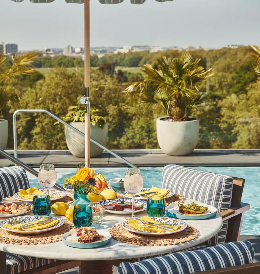 Poolside dining table with striped chairs, colourful plates, and yellow napkins under blue-and-white umbrella.