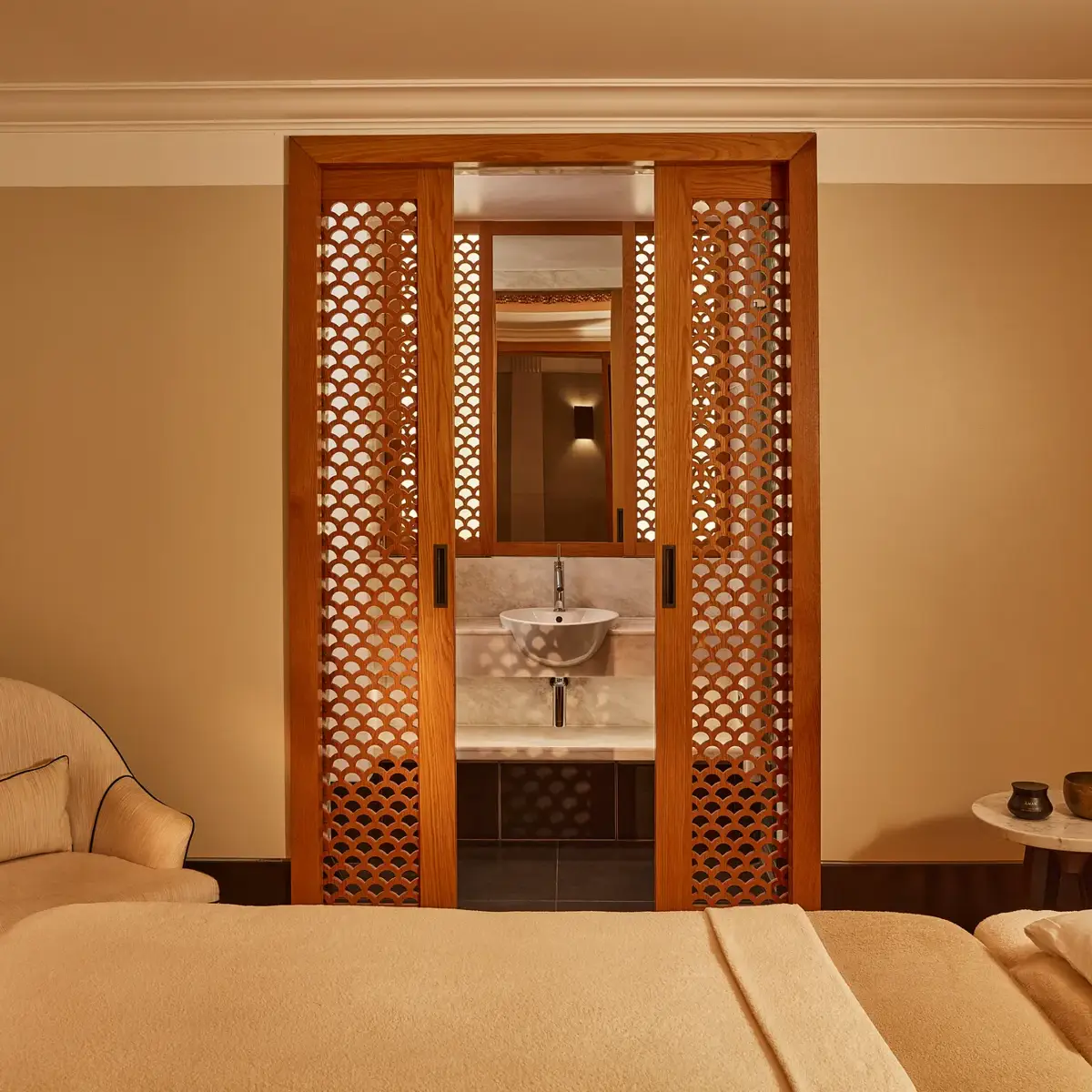 Tranquil spa room with a treatment bed, soft armchair, and carved wooden screens revealing a small sink area.