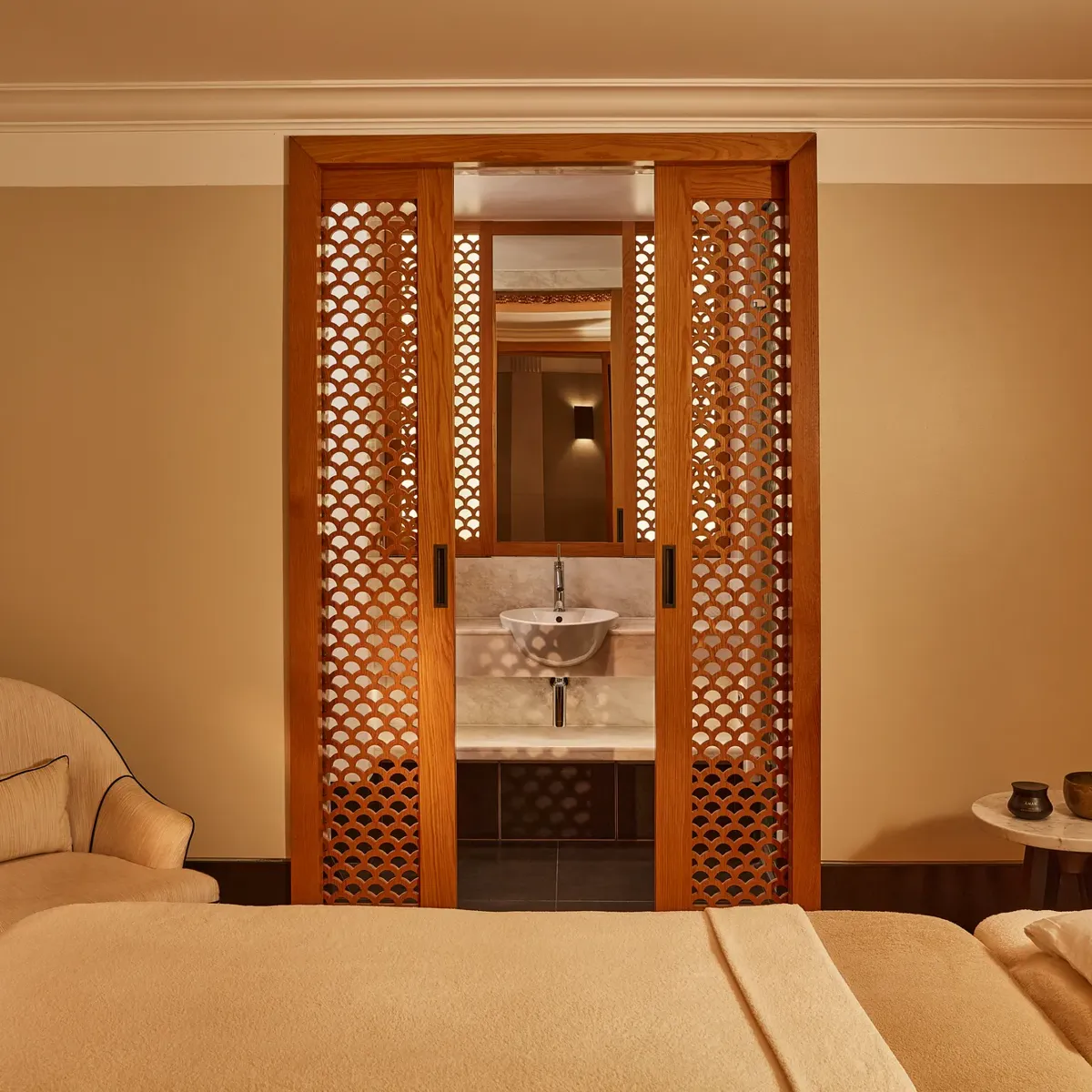 Tranquil spa room with a treatment bed, soft armchair, and carved wooden screens revealing a small sink area.