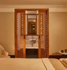 Tranquil spa room with a treatment bed, soft armchair, and carved wooden screens revealing a small sink area.