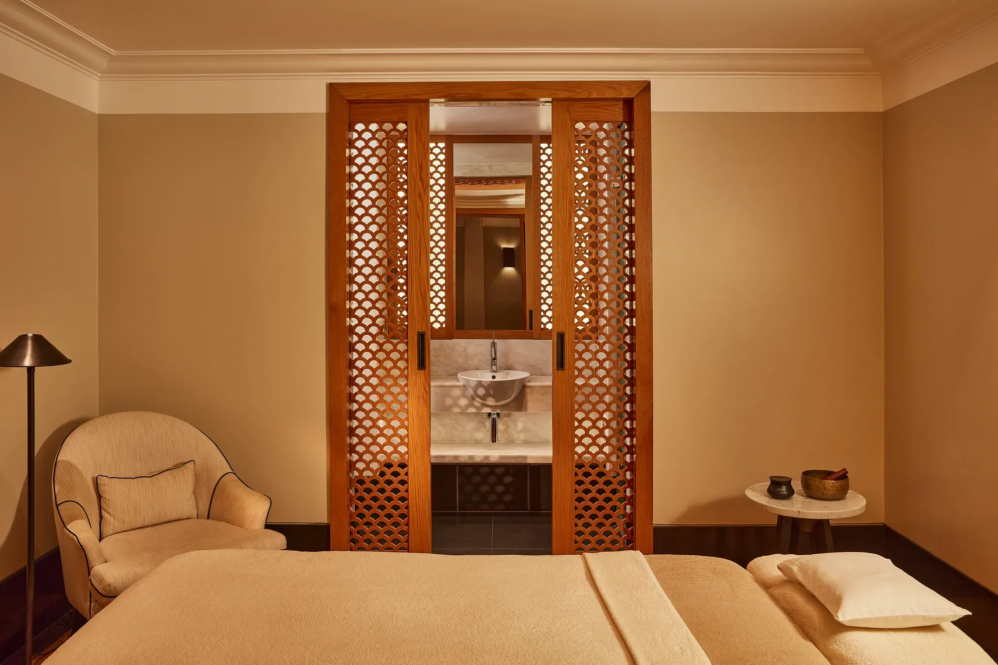 Tranquil spa room with a treatment bed, soft armchair, and carved wooden screens revealing a small sink area.