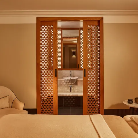 Tranquil spa room with a treatment bed, soft armchair, and carved wooden screens revealing a small sink area.