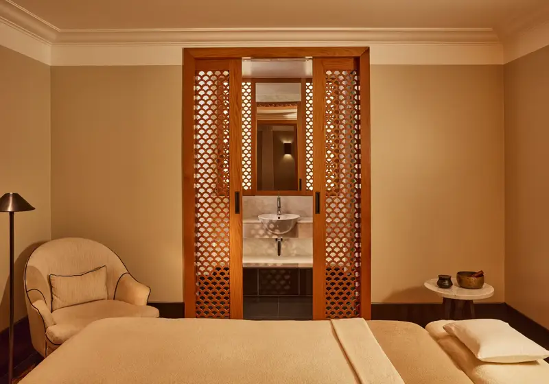 Tranquil spa room with a treatment bed, soft armchair, and carved wooden screens revealing a small sink area.