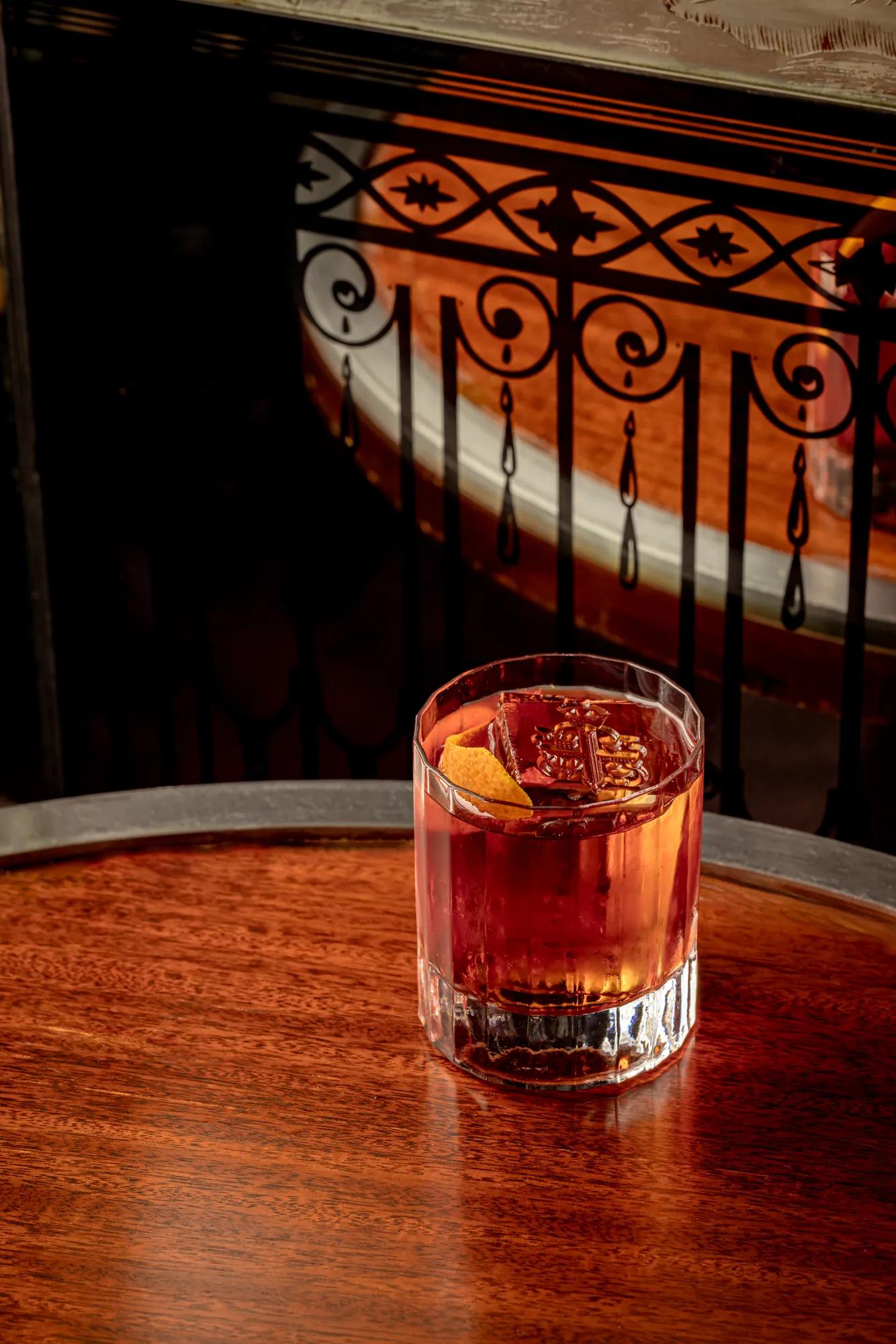 Short glass of amber cocktail with ice and orange peel on wooden table, set against ornate black railing and warm light.