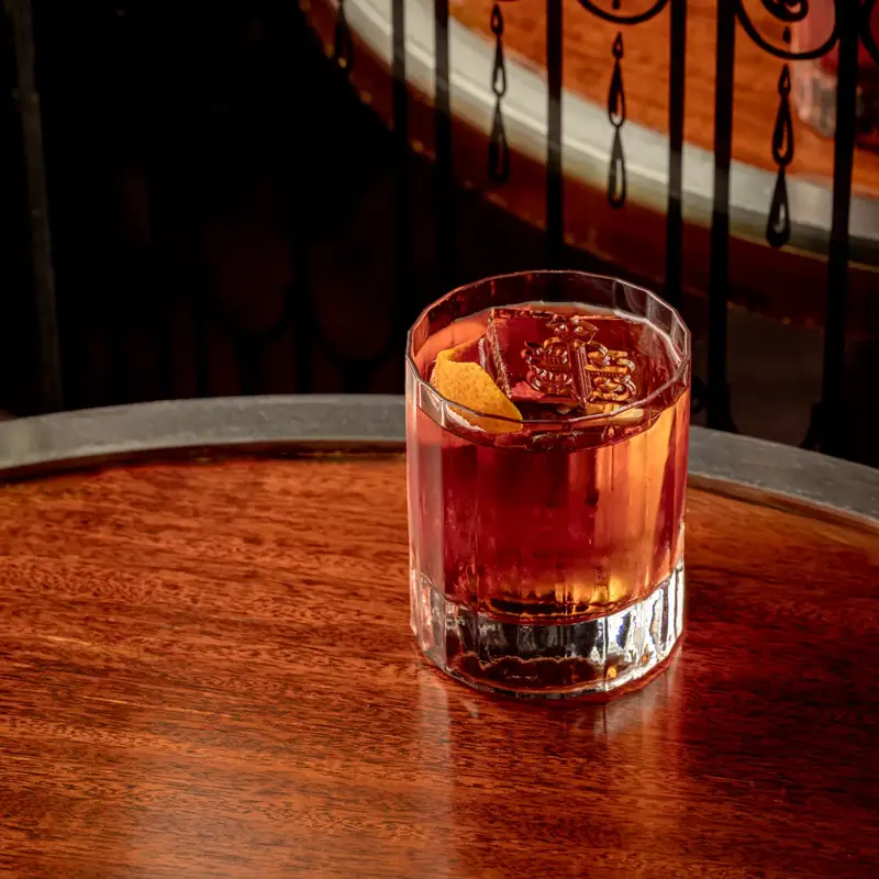 Short glass of amber cocktail with ice and orange peel on wooden table, set against ornate black railing and warm light.