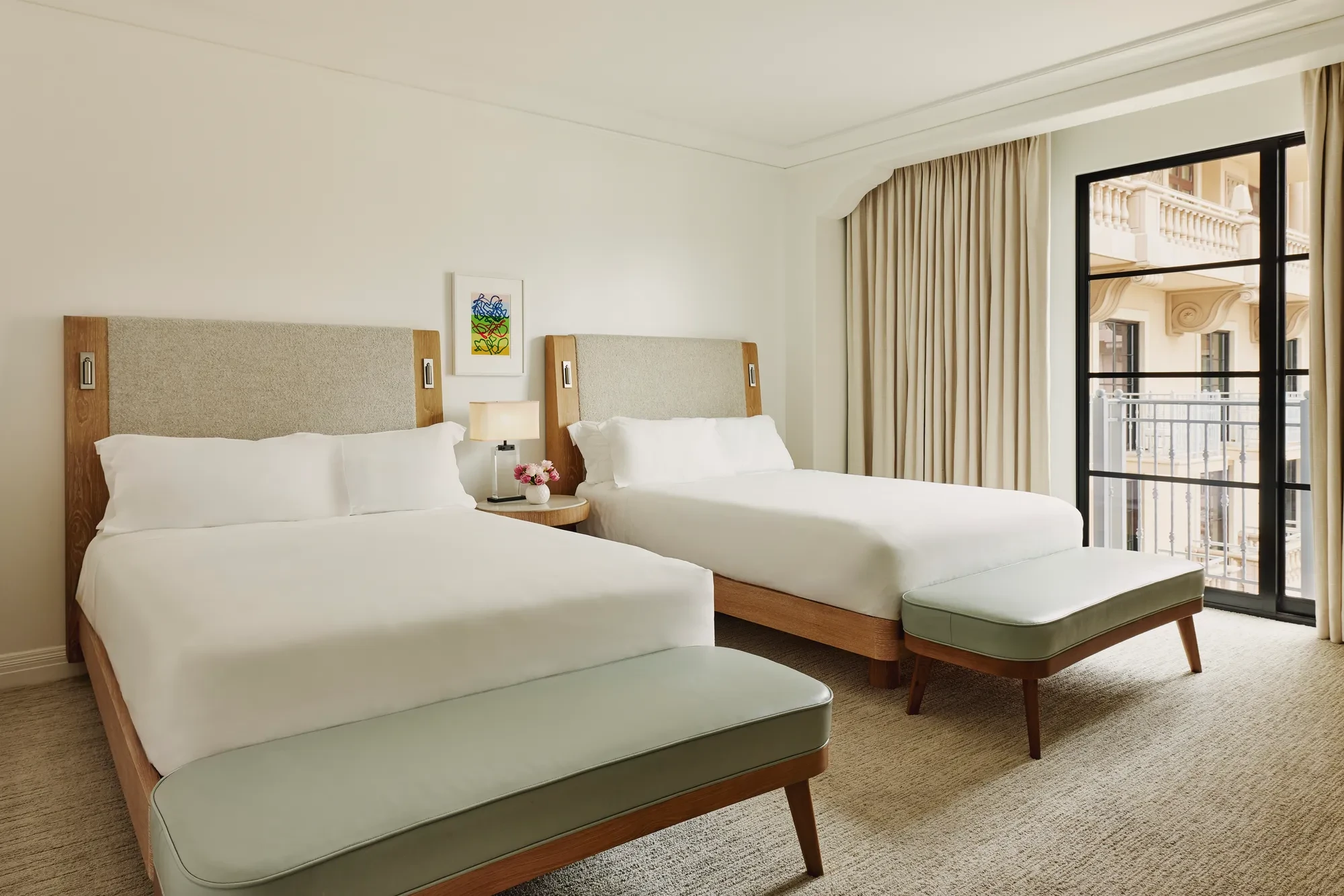 Two-queen bedroom with upholstered headboards, benches at each bed, nightstand with lamp and flowers, and balcony doors with curtains.
