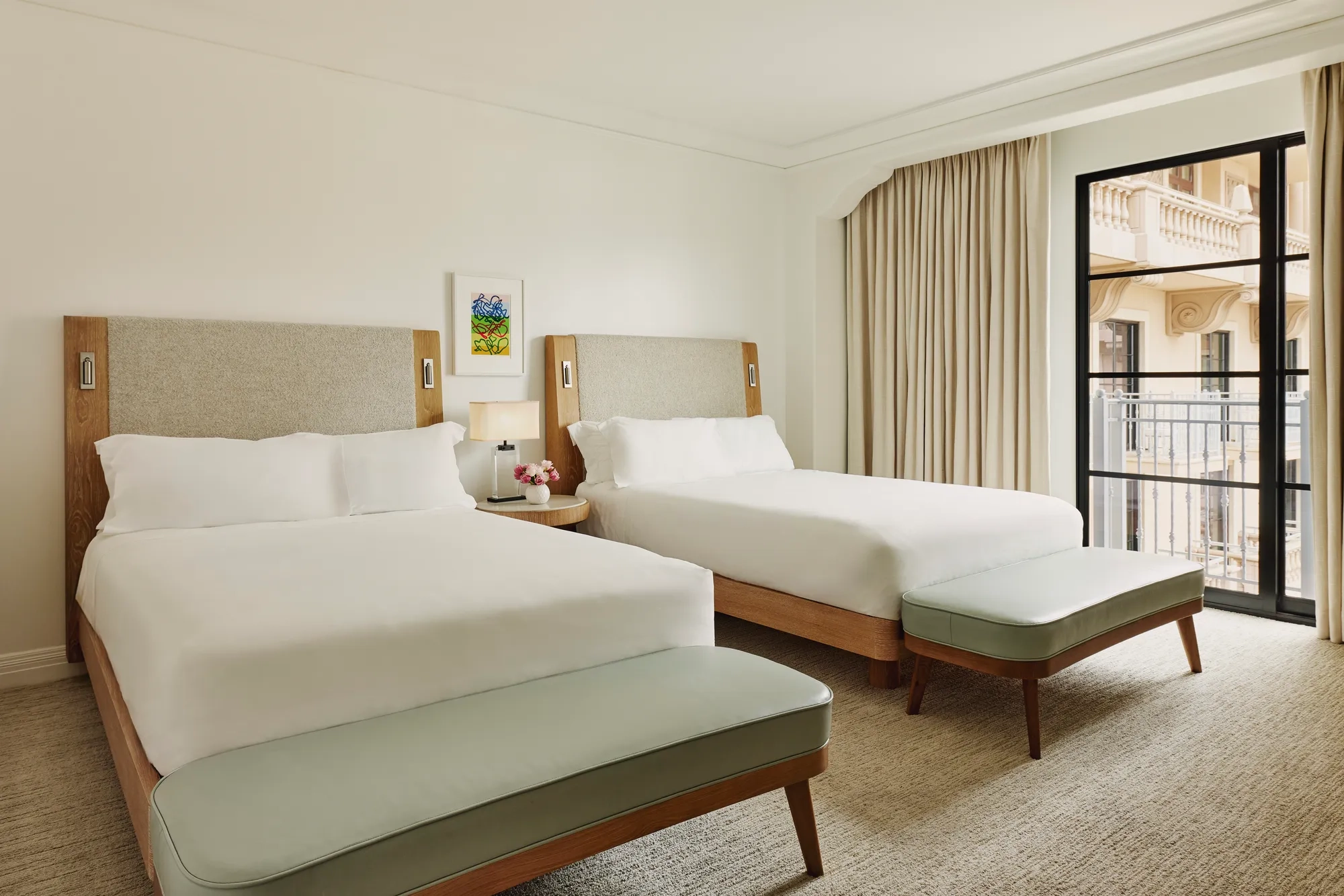 Two-queen bedroom with upholstered headboards, benches at each bed, nightstand with lamp and flowers, and balcony doors with curtains.