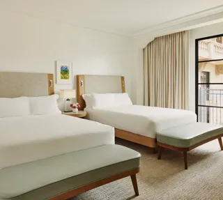 Two-queen bedroom with upholstered headboards, benches at each bed, nightstand with lamp and flowers, and balcony doors with curtains.