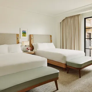 Two-queen bedroom with upholstered headboards, benches at each bed, nightstand with lamp and flowers, and balcony doors with curtains.