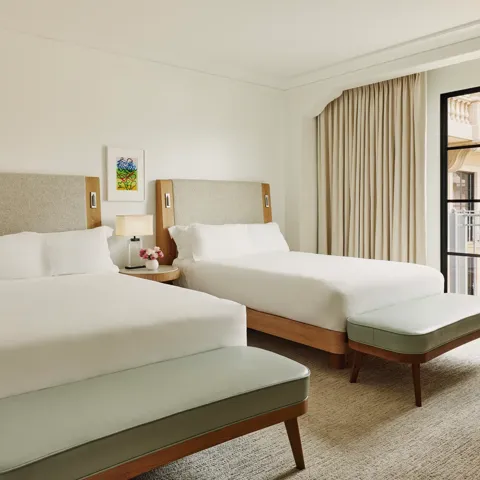 Two-queen bedroom with upholstered headboards, benches at each bed, nightstand with lamp and flowers, and balcony doors with curtains.