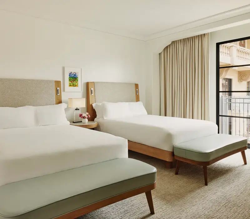 Two-queen bedroom with upholstered headboards, benches at each bed, nightstand with lamp and flowers, and balcony doors with curtains.