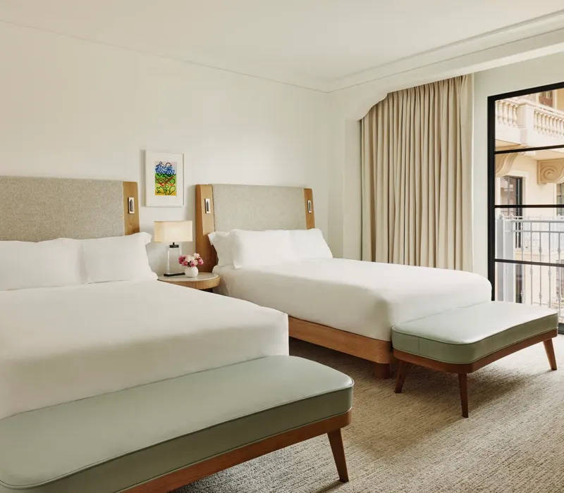 Two-queen bedroom with upholstered headboards, benches at each bed, nightstand with lamp and flowers, and balcony doors with curtains.