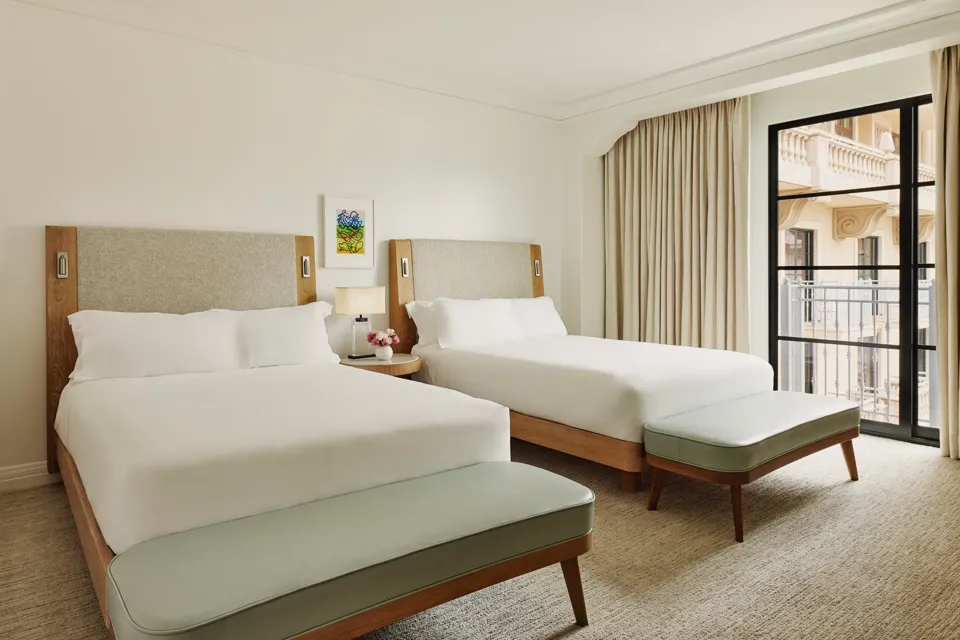 Two-queen bedroom with upholstered headboards, benches at each bed, nightstand with lamp and flowers, and balcony doors with curtains.