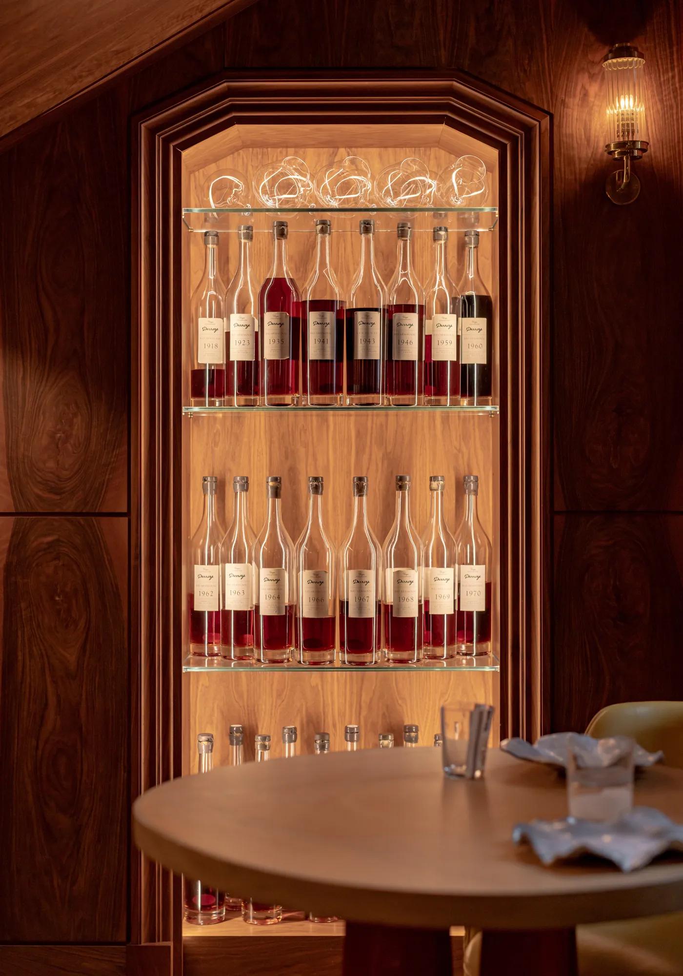 Backlit display of wine and spirits bottles on glass shelves within a wood-paneled cabinet, with a table set in the foreground