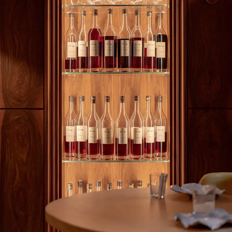 Backlit display of wine and spirits bottles on glass shelves within a wood-paneled cabinet, with a table set in the foreground