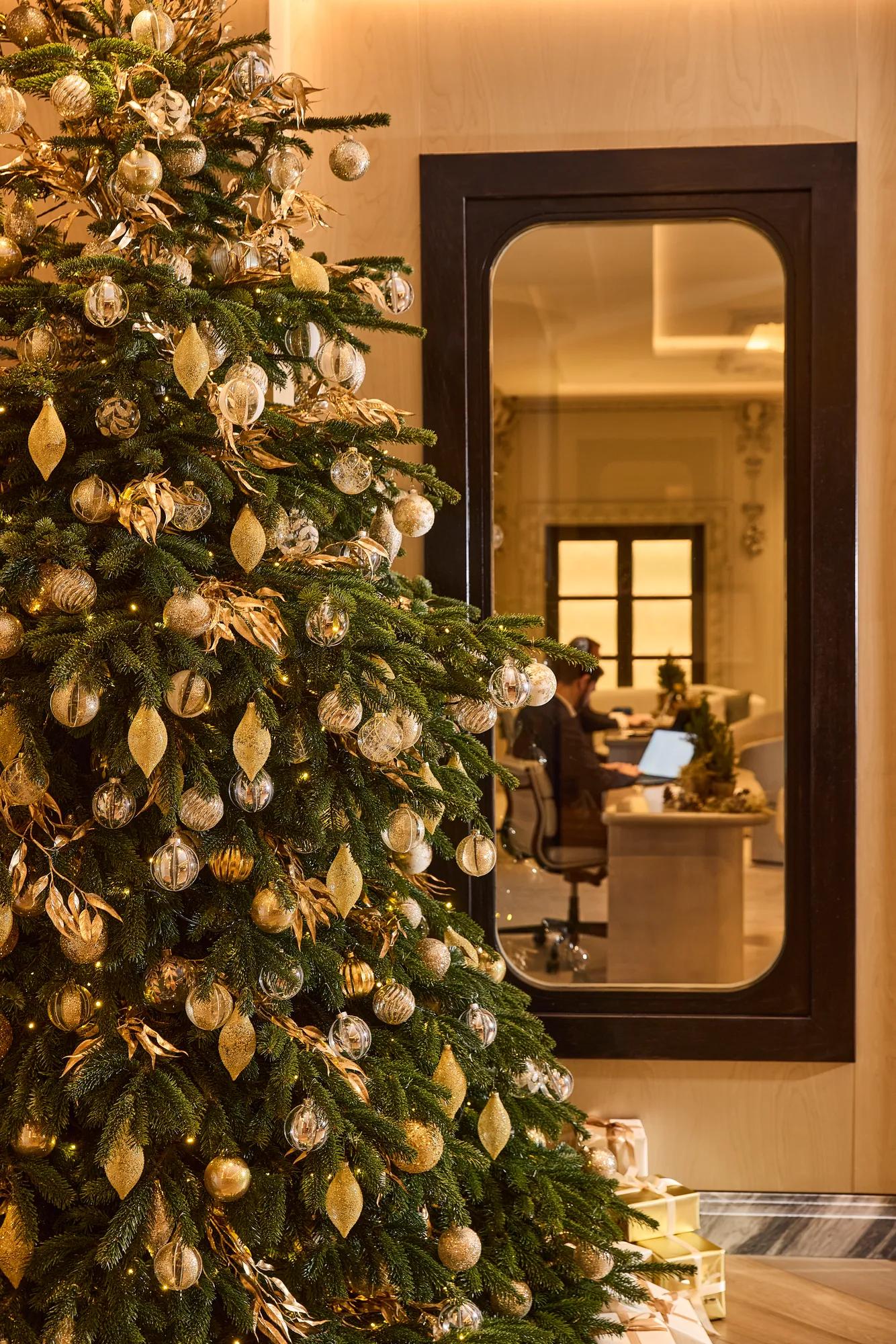 A beautifully decorated Christmas tree adorned with gold, silver and glass ornaments stands in an elegant hotel interior. Reflected in a large framed mirror, a reception area can be seen in the background, while wrapped gifts at the base of the tree add a festive touch.