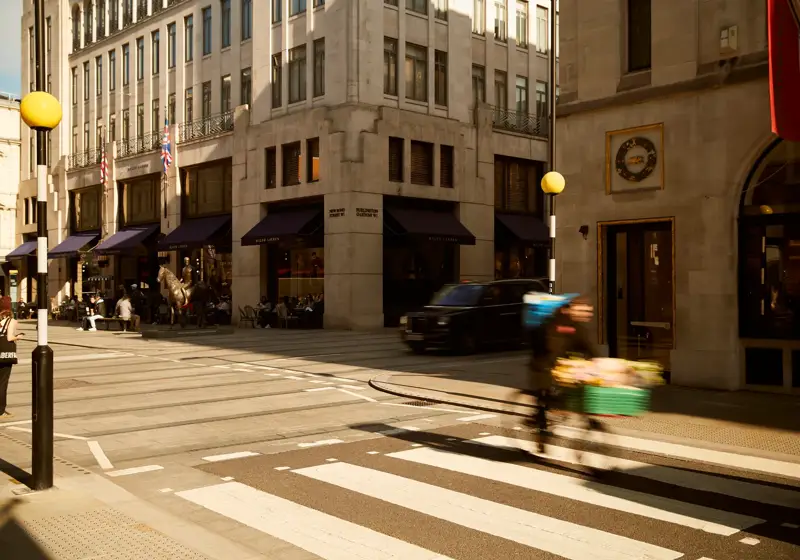 A busy London street corner with a cyclist crossing a zebra crossing in motion blur, a black cab driving by, and people walking near shops with navy awnings under a cream stone building.