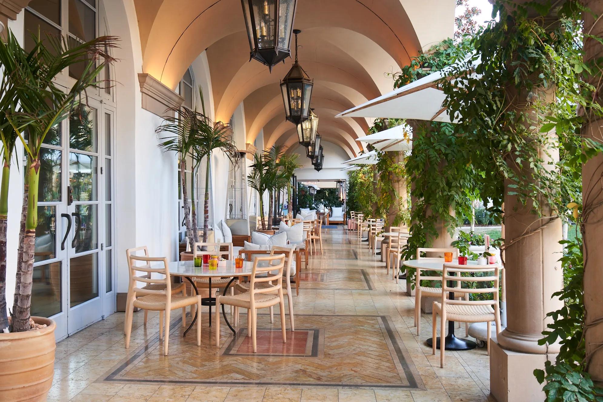 Open-air terrace with arched ceilings, lush greenery, white umbrellas, and elegant café seating along a sunlit colonnade.