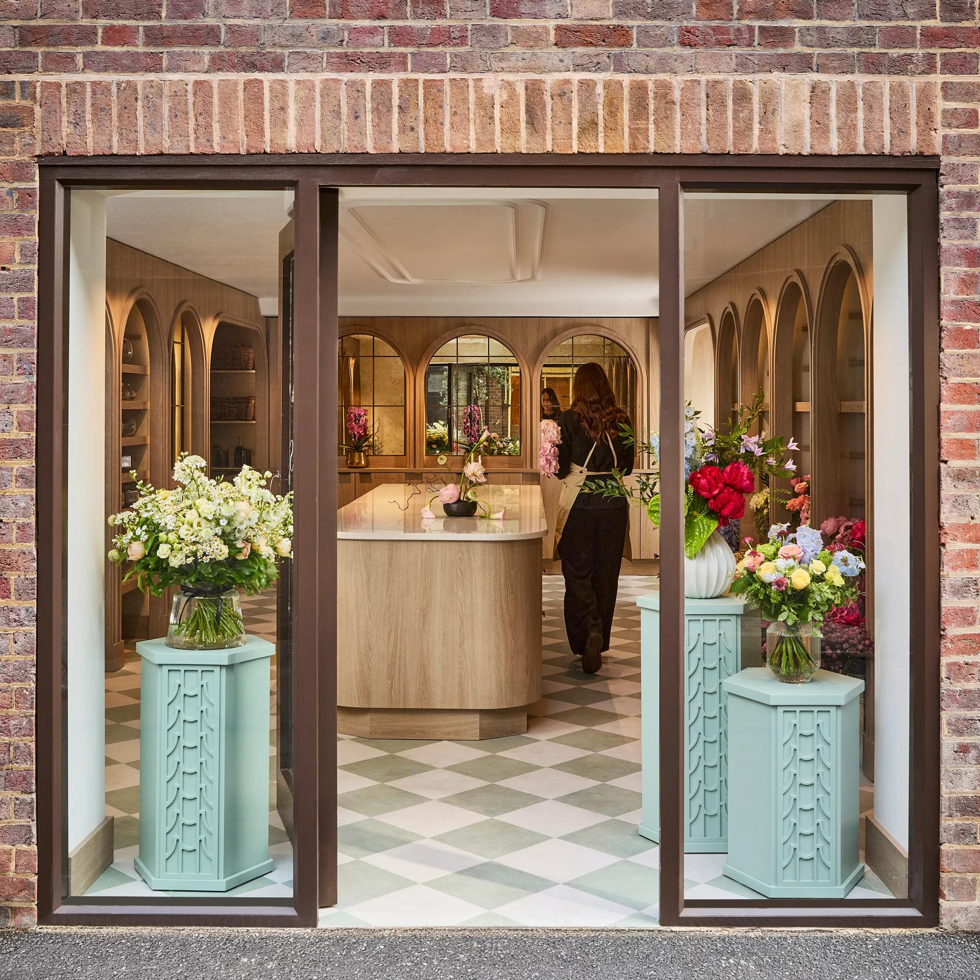 **Alt text (WCAG 2.2 AA, under 125 characters):**

View through a boutique window into a florist, with fresh flower displays, oak interiors and a florist arranging blooms.
