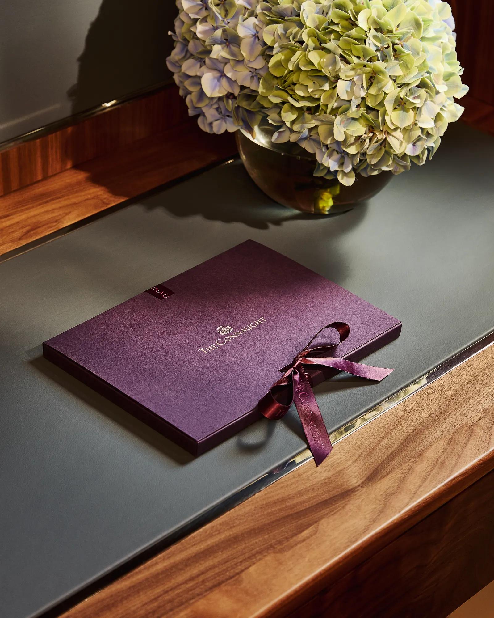The Connaught welcome booklet with ribbon placed on a desk beside a vase of hydrangeas in a softly lit hotel room.