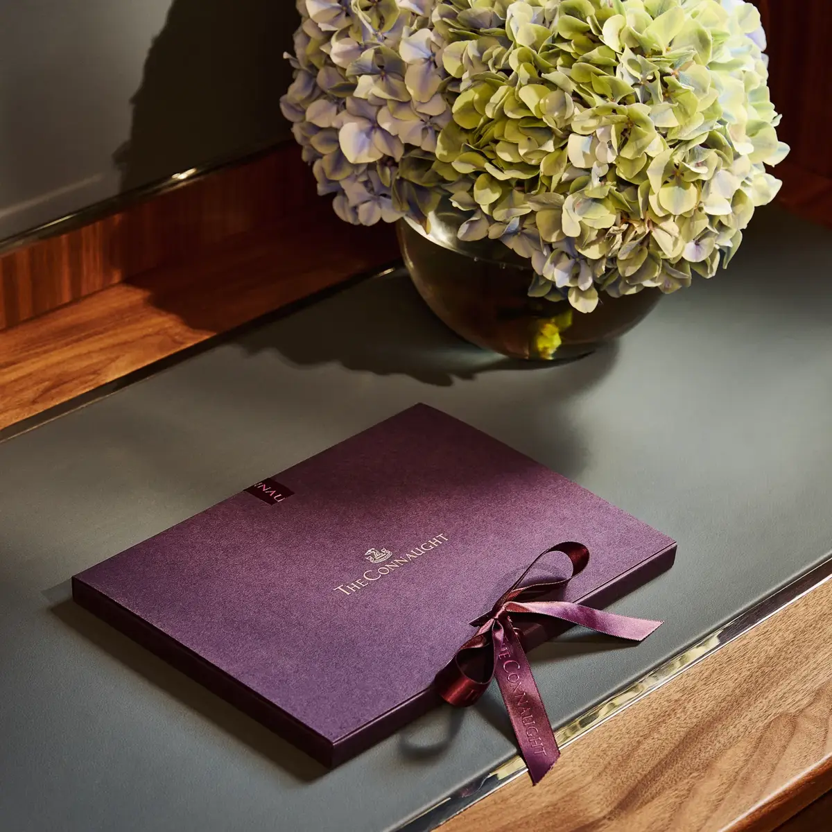 The Connaught welcome booklet with ribbon placed on a desk beside a vase of hydrangeas in a softly lit hotel room.