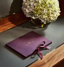 The Connaught welcome booklet with ribbon placed on a desk beside a vase of hydrangeas in a softly lit hotel room.