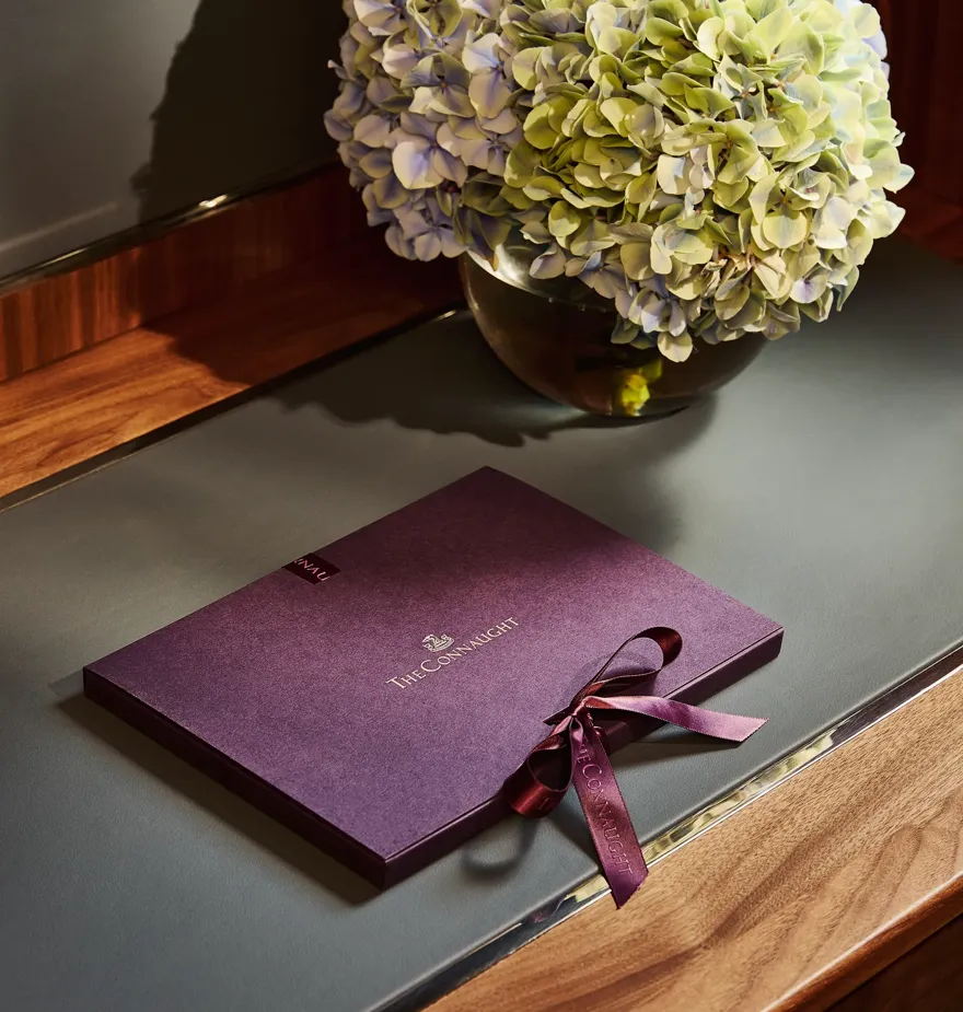 The Connaught welcome booklet with ribbon placed on a desk beside a vase of hydrangeas in a softly lit hotel room.