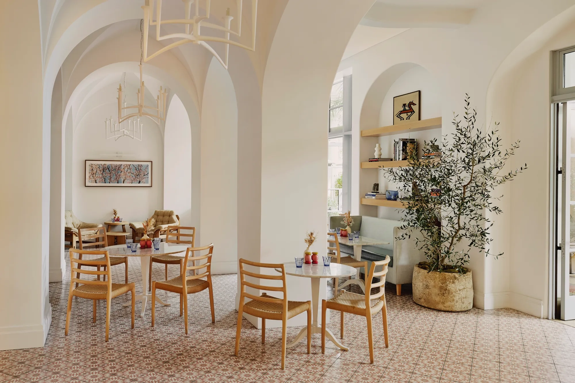 Light café interior with vaulted arches, patterned tile floor, pale wood chairs, and a potted olive tree.