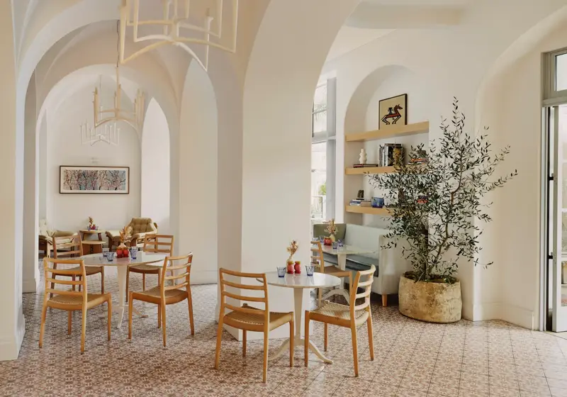 Light café interior with vaulted arches, patterned tile floor, pale wood chairs, and a potted olive tree.