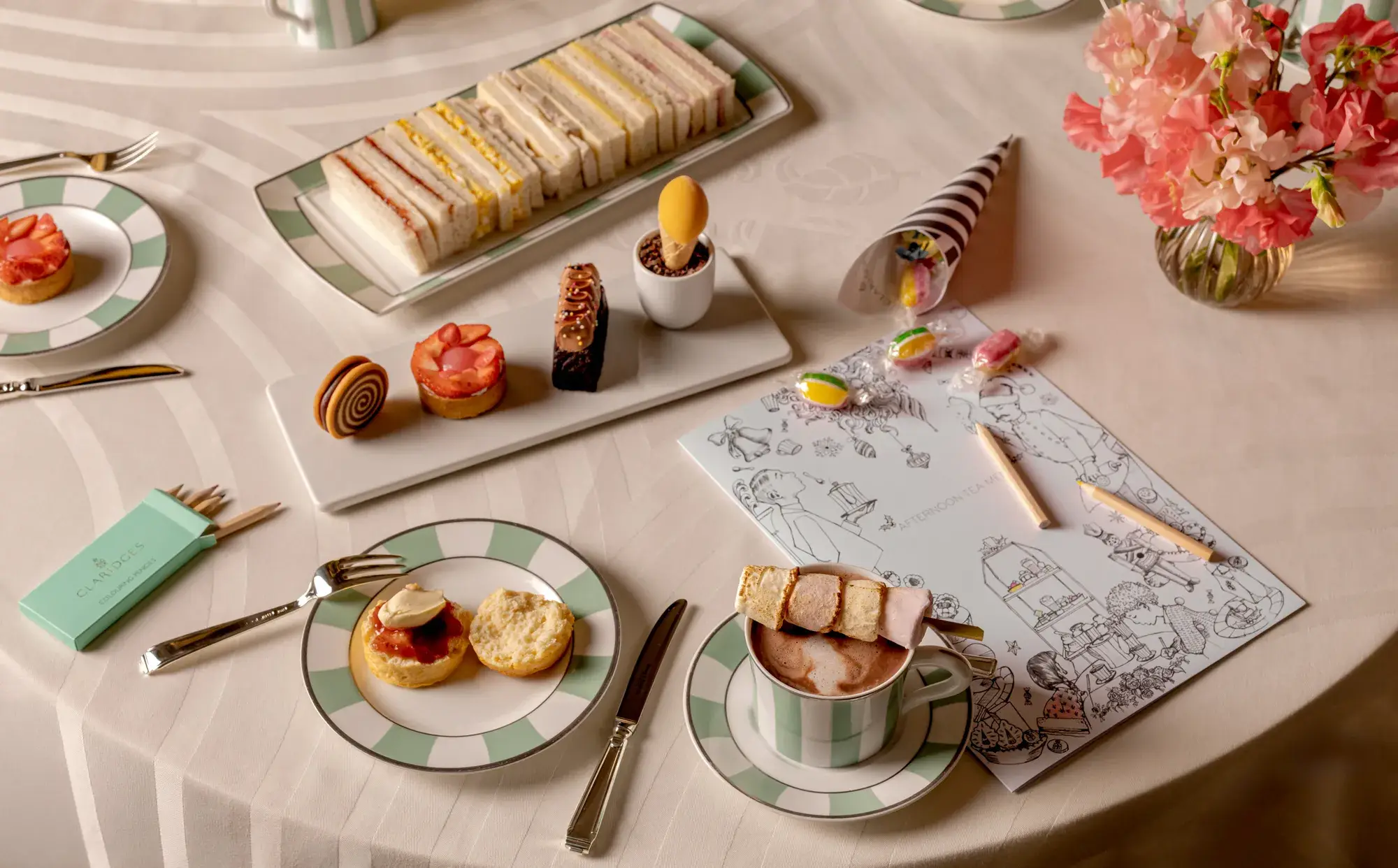 Children’s afternoon tea with finger sandwiches, pastries, marshmallow hot chocolate and colouring sheet on table.