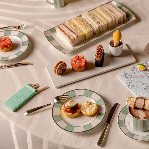 Children’s afternoon tea with finger sandwiches, pastries, marshmallow hot chocolate and colouring sheet on table.
