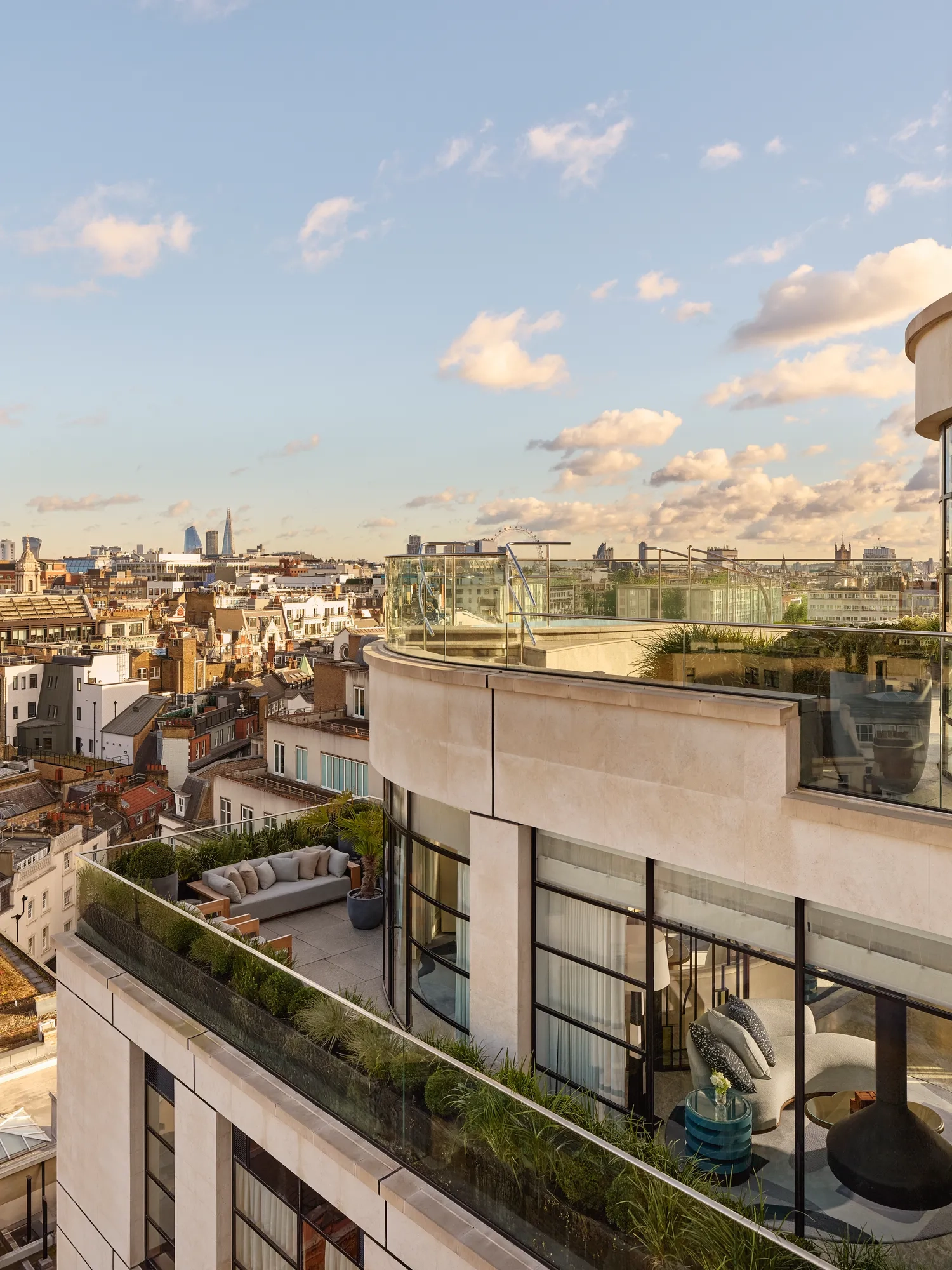 Exterior view of a luxury penthouse with wraparound terraces featuring outdoor seating, greenery, and glass balustrades, overlooking London’s skyline under a clear evening sky.
